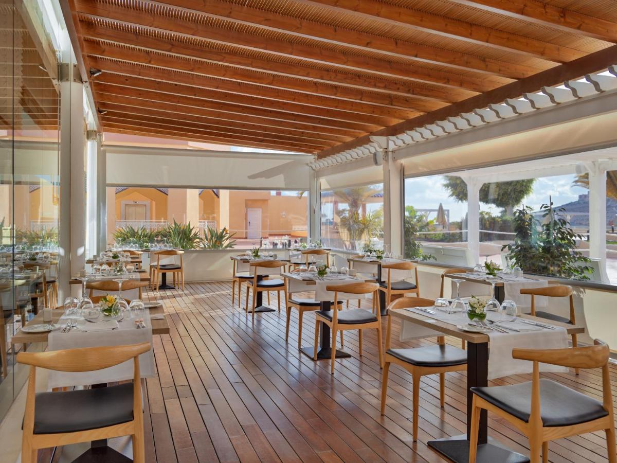 Un restaurante con mesas y sillas blancas y ventanas. en H10 Playa Esmeralda - Adults Only, en Costa Calma