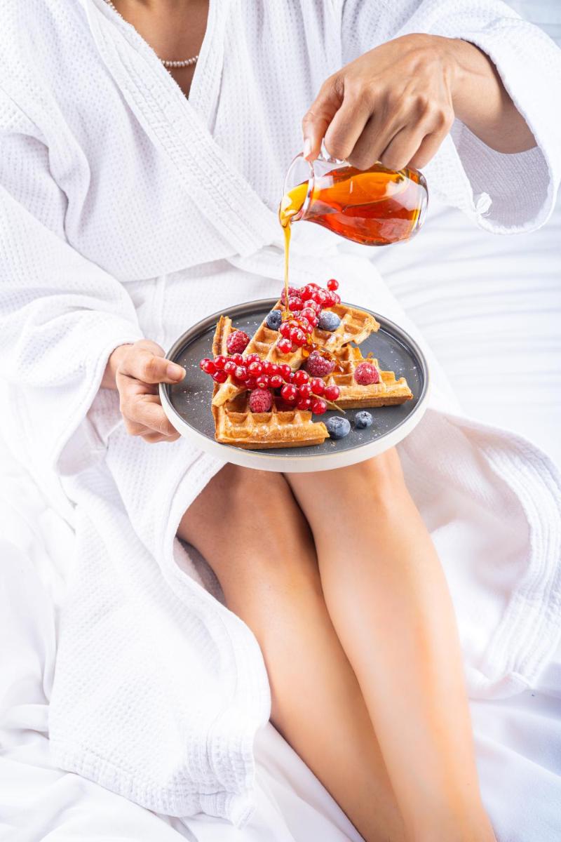 a woman sitting in bed holding a plate of waffles and syrup at Hyatt Regency Dehradun Resort and Spa in Dehradun