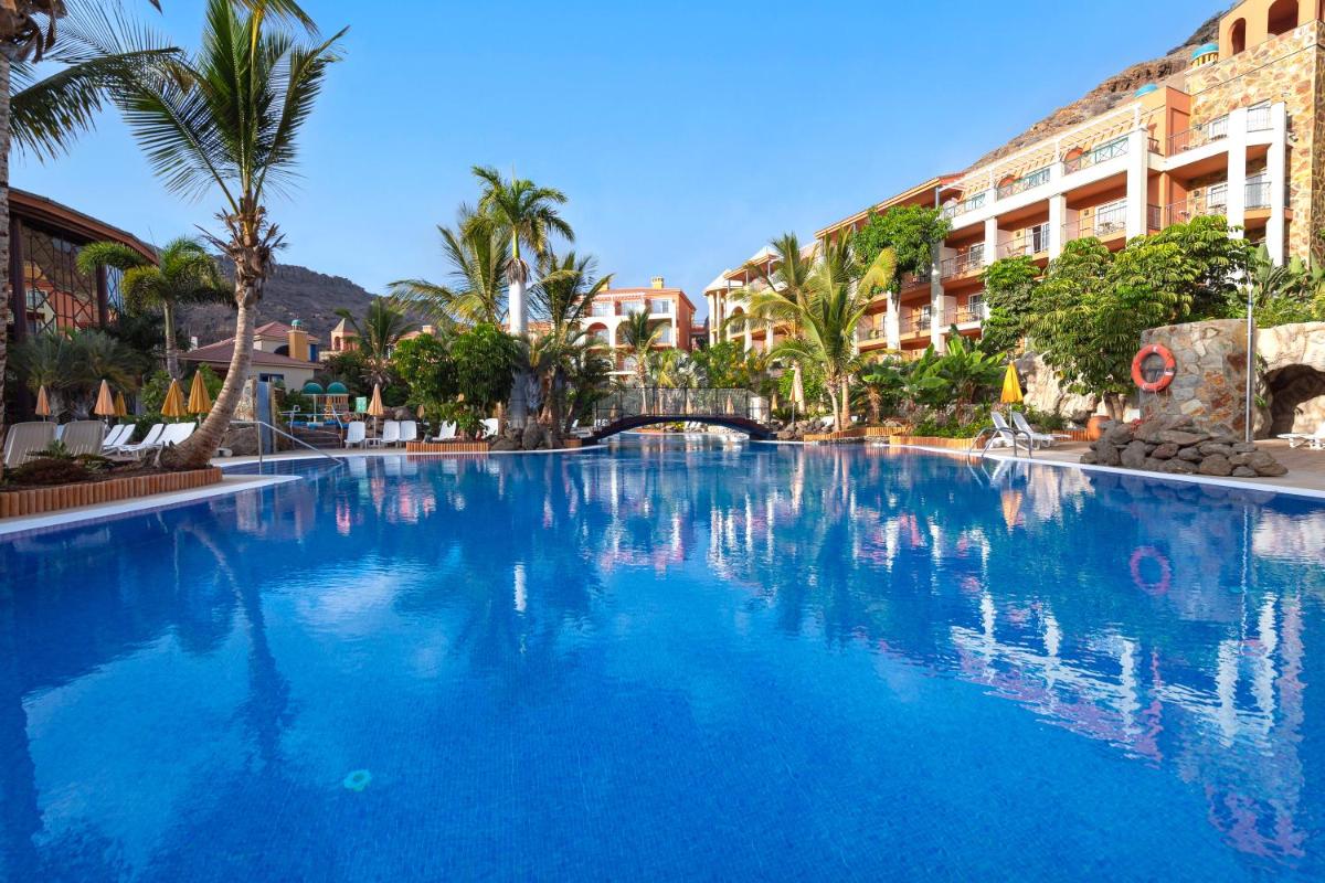 Una gran piscina azul con palmeras y edificios. en Hotel Cordial Mogán Playa, en Puerto de Mogán