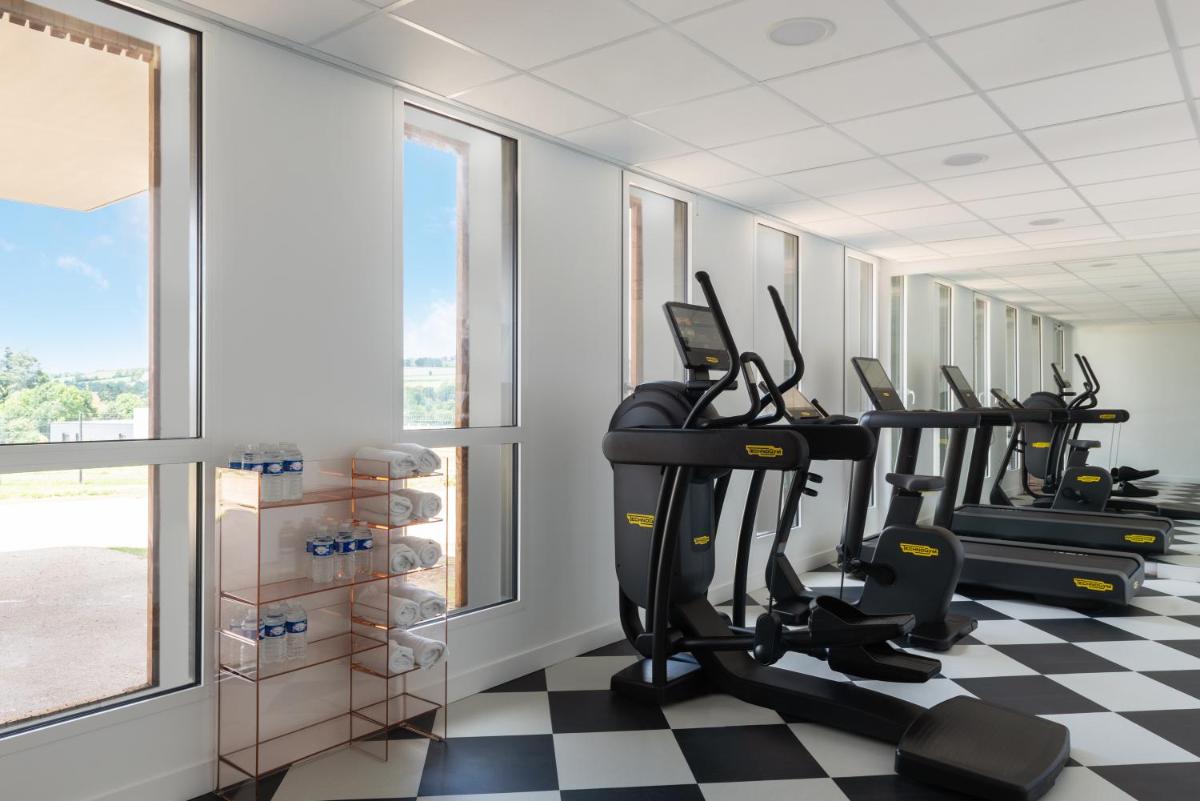 une salle de sport avec tapis de course et vélos elliptiques dans une pièce avec fenêtres dans l'établissement Hôtel Omnubo Collection, aux Épesses
