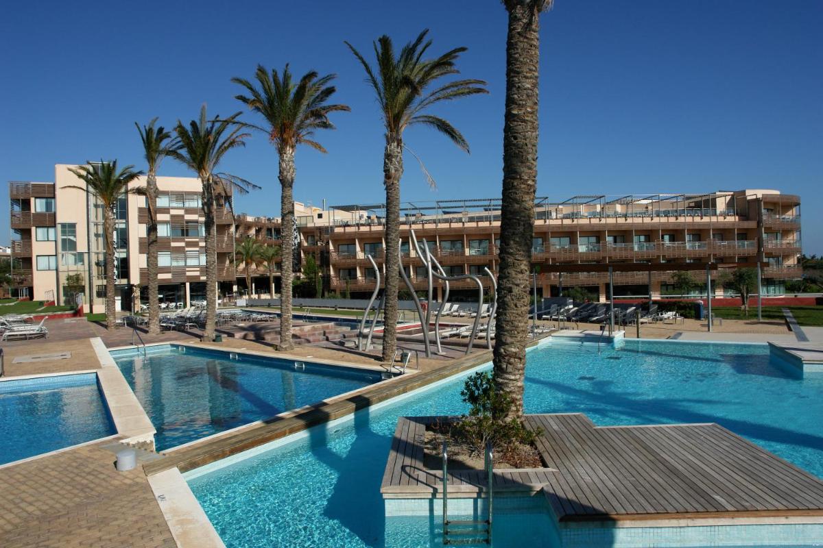 Una piscina con palmeras y un edificio. en Ohtels Les Oliveres, en Perelló