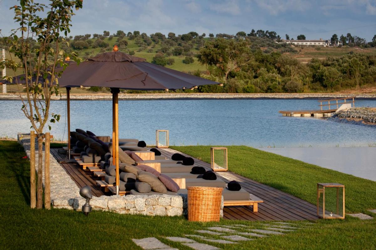 um grupo de bancos com um guarda-chuva ao lado de um lago em L'AND Vineyards - Relais & Châteaux em Montemor-o-Novo