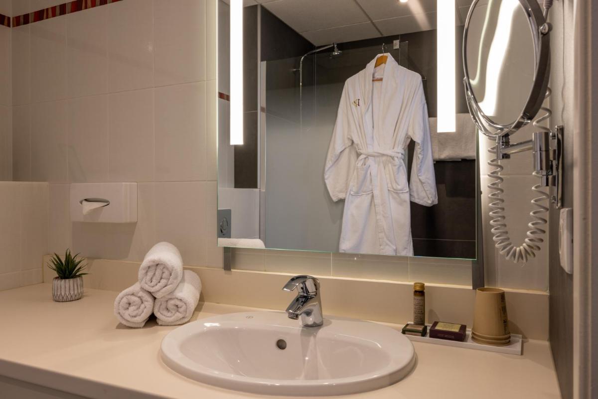 La salle de bains est pourvue d'un lavabo et d'un peignoir blanc suspendu dans un miroir. dans l'établissement Hôtel Val-Vignes Colmar Haut-Koenigsbourg, The Originals Relais, à Saint-Hippolyte