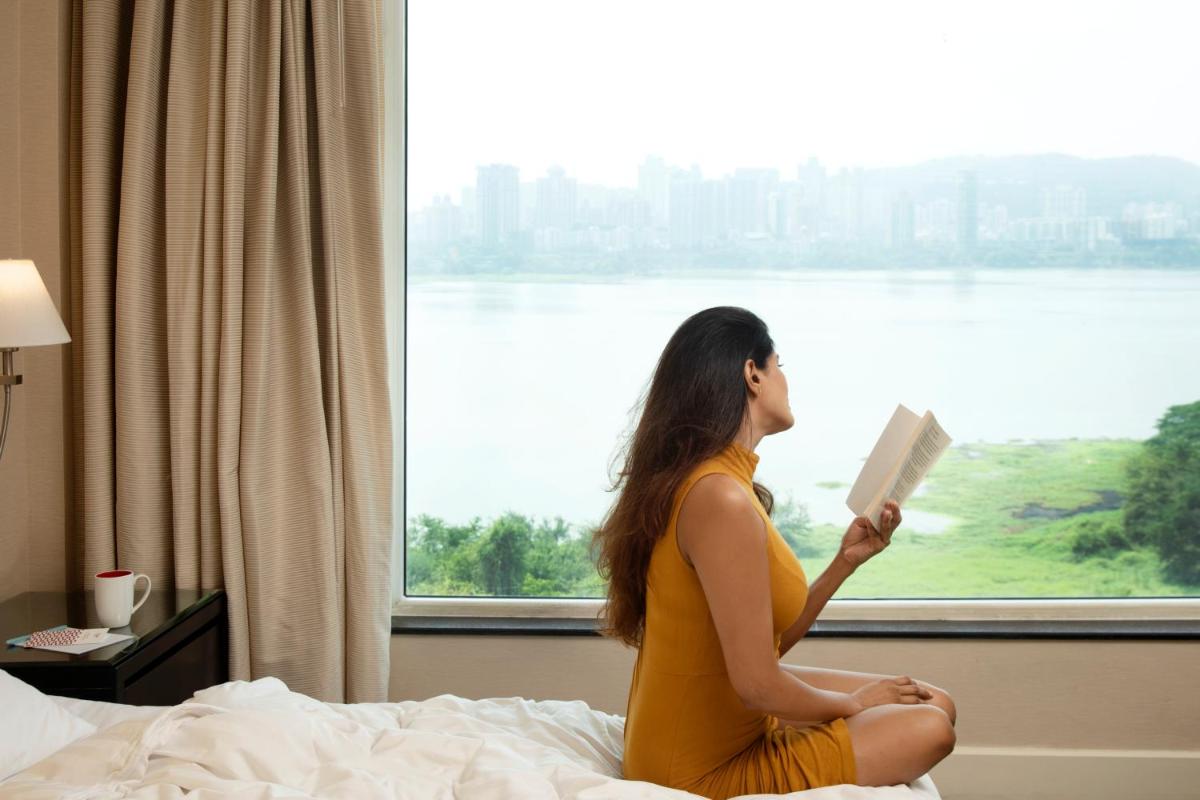 a woman sitting on a bed reading a book in front of a window at Marriott Executive Apartment - Lakeside Chalet, Mumbai in Mumbai