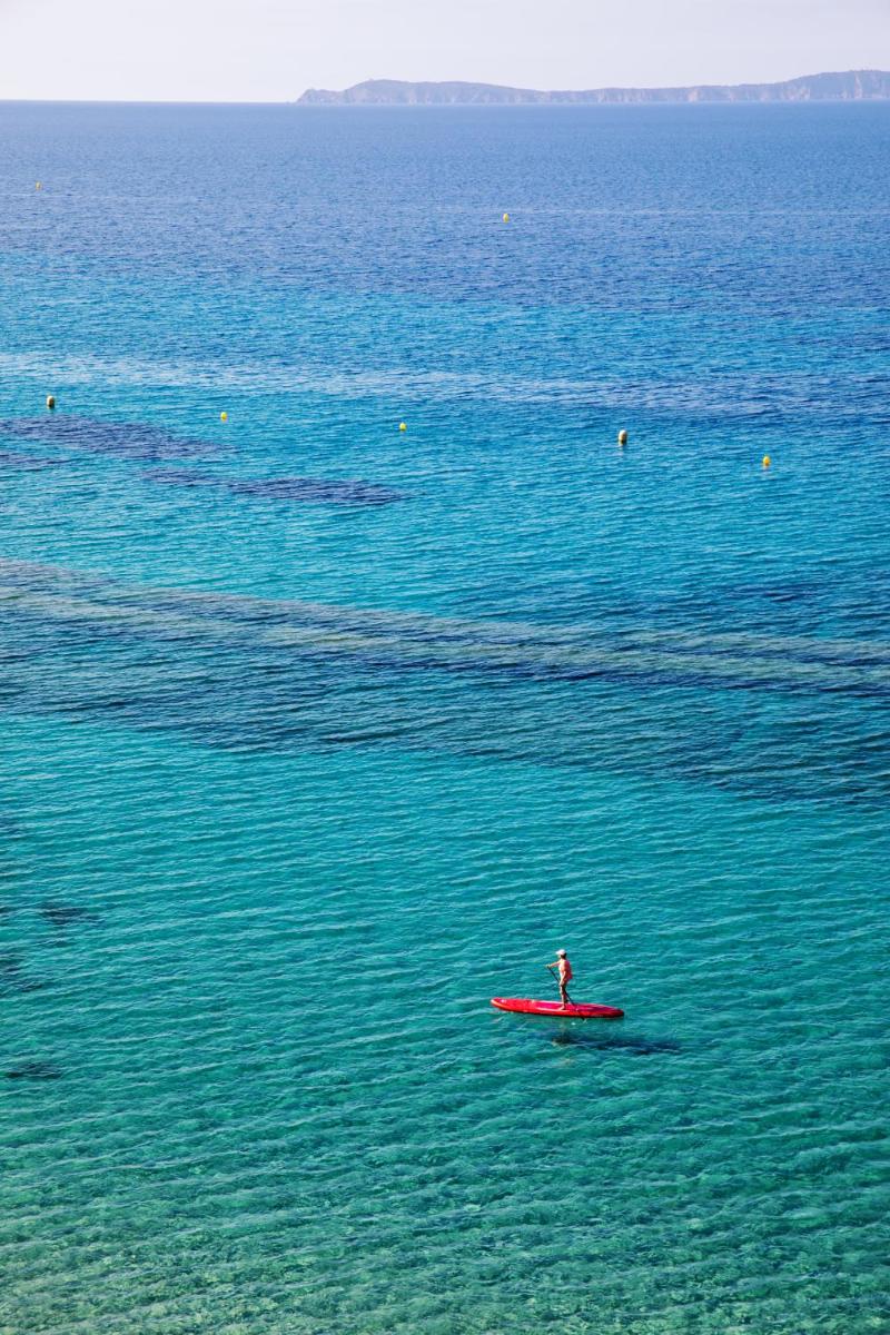 une personne assise sur une planche de surf rouge dans l'eau dans l'établissement Hôtel Le Bailli De Suffren, à Rayol-Canadel-sur-Mer