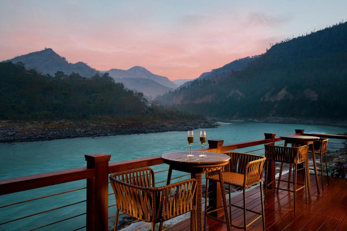 a table and chairs on a deck with a view of a river at Anand Kashi by the Ganges Rishikesh - IHCL Seleqtions in Rishīkesh