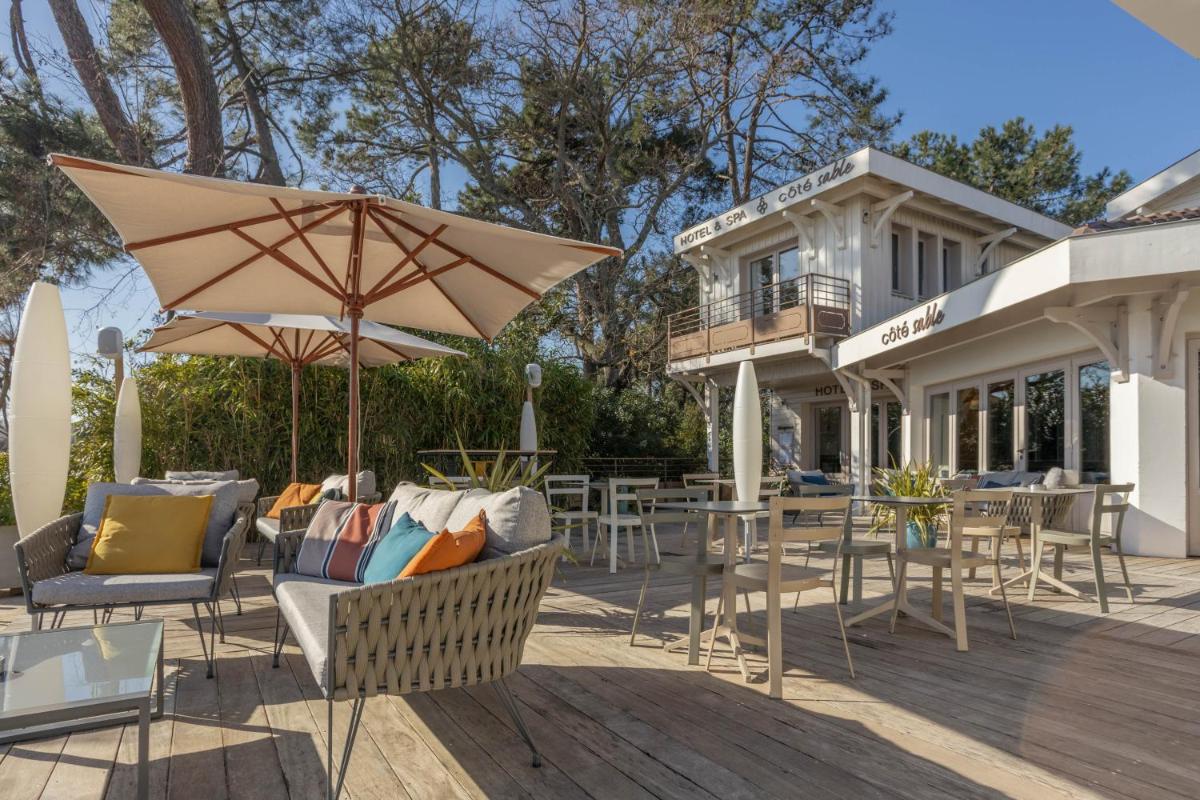 une terrasse avec des chaises, des tables et un parasol dans l'établissement Hôtel Côté Sable, à Cap-Ferret