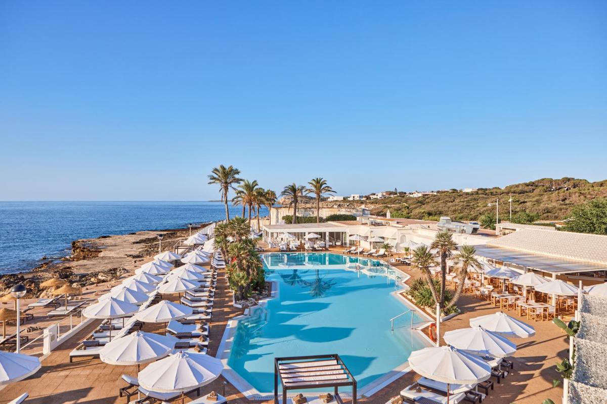 Una piscina con sombrillas y sillas y el océano. en AluaSoul Menorca - Adults Only, en S'Algar