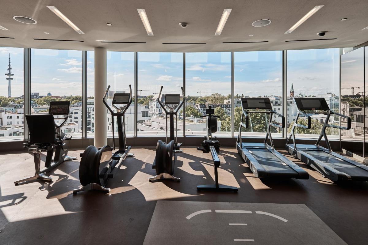 einen Fitnessraum mit Kardiogeräten in einem Gebäude mit Fenstern in der Unterkunft The Fontenay Hamburg in Hamburg einen Fitnessraum mit Kardiogeräten in einem Gebäude mit Fenstern in der Unterkunft The Fontenay Hamburg in Hamburg