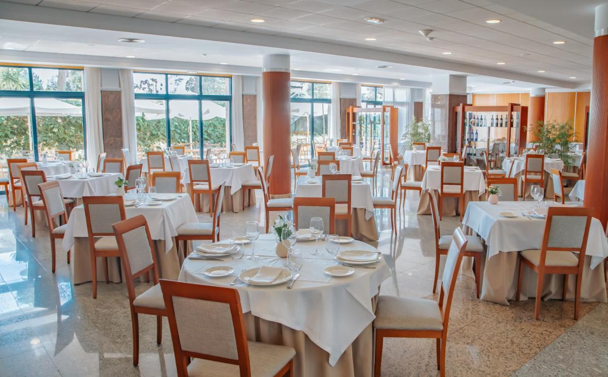 Un comedor con mesas y sillas blancas y ventanas. en Hotel Spa Galatea, en Sanxenxo