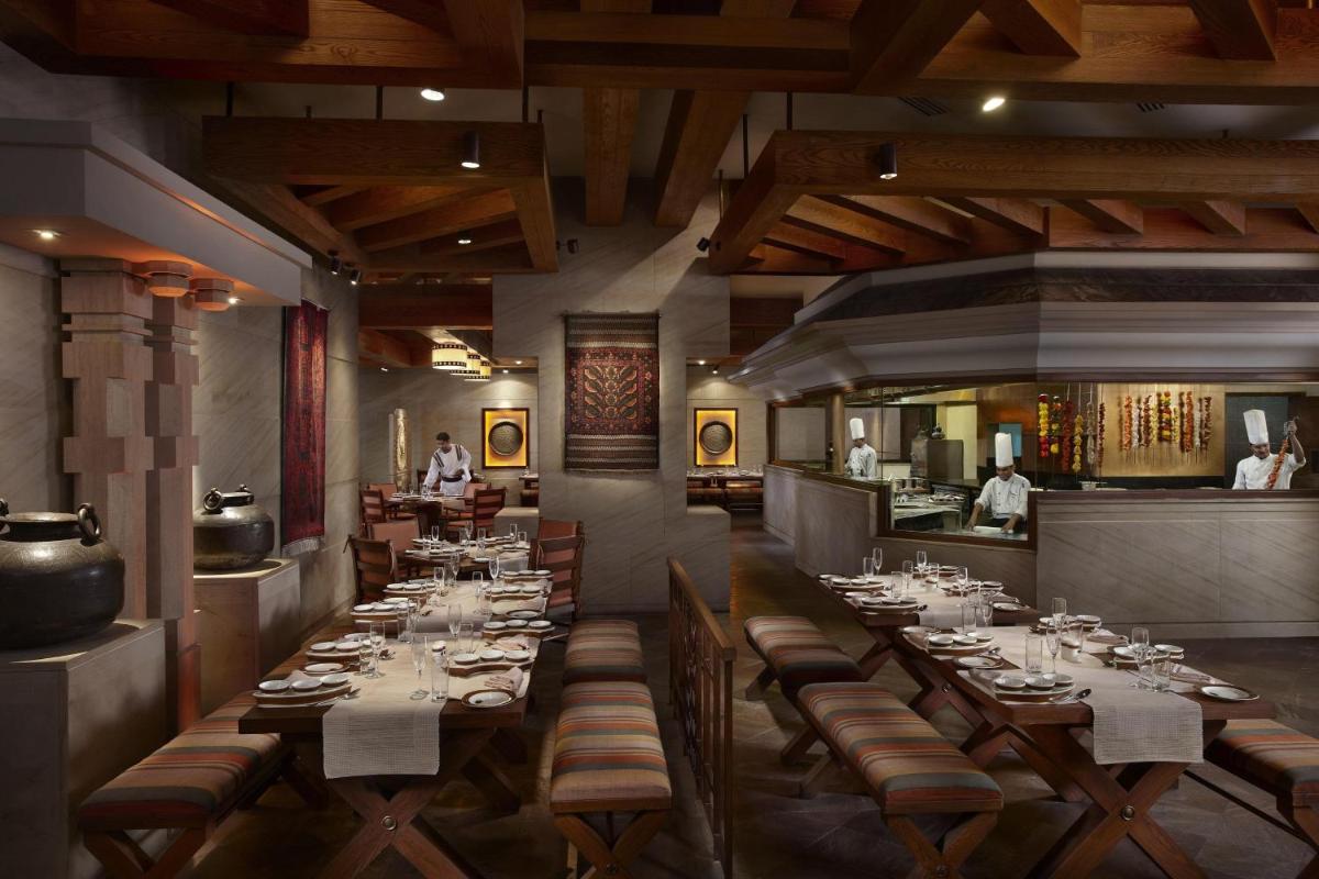 a dining room with long tables and people in the kitchen at ITC Gardenia, a Luxury Collection Hotel, Bengaluru in Bengaluru a dining room with long tables and people in the kitchen at ITC Gardenia, a Luxury Collection Hotel, Bengaluru in Bengaluru