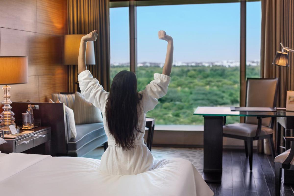 a woman is standing on a bed looking out a window at The Leela Ambience Gurugram Hotel & Residences - The Luxury Urban Sanctuary in Gurgaon