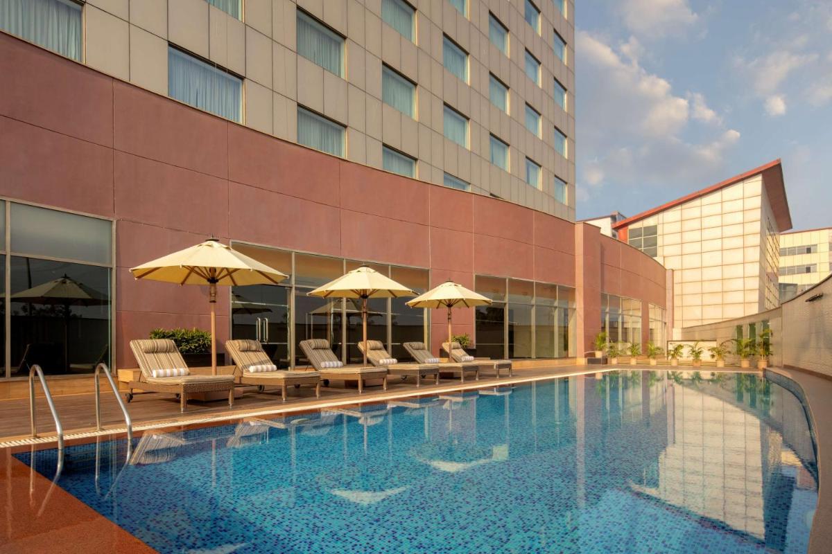 a swimming pool with chairs and umbrellas next to a building at Radisson Gurugram Udyog Vihar in Gurgaon a swimming pool with chairs and umbrellas next to a building at Radisson Gurugram Udyog Vihar in Gurgaon