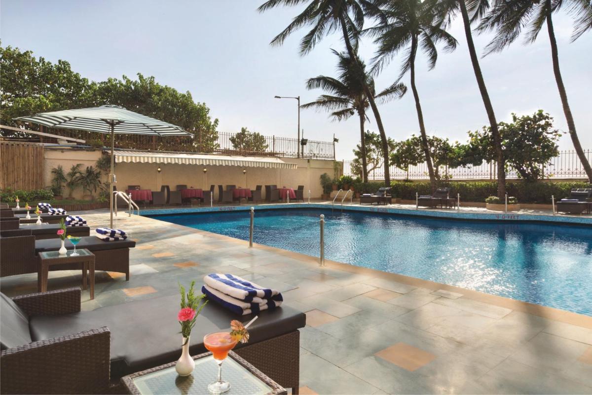 a swimming pool with tables and chairs and palm trees at Ramada Plaza By Wyndham Palm Grove in Mumbai a swimming pool with tables and chairs and palm trees at Ramada Plaza By Wyndham Palm Grove in Mumbai