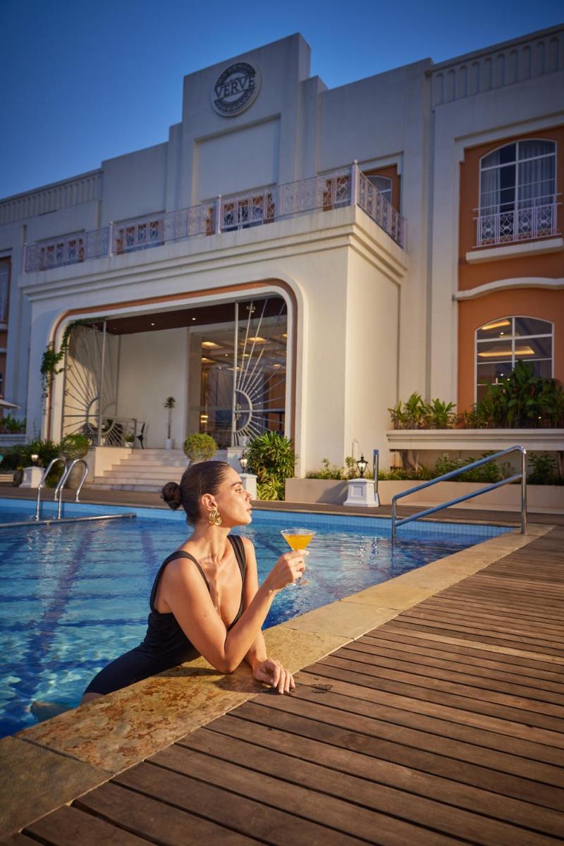 a woman sitting next to a swimming pool with a drink at Miros - Formerly Imperial Goa in Old Goa