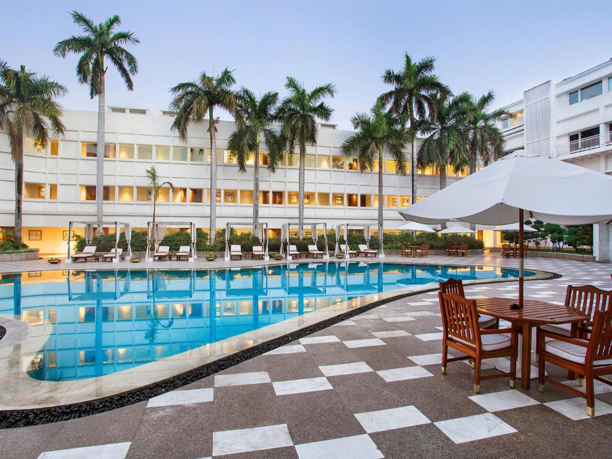 a hotel pool with tables and chairs and palm trees at The Claridges New Delhi in New Delhi a hotel pool with tables and chairs and palm trees at The Claridges New Delhi in New Delhi