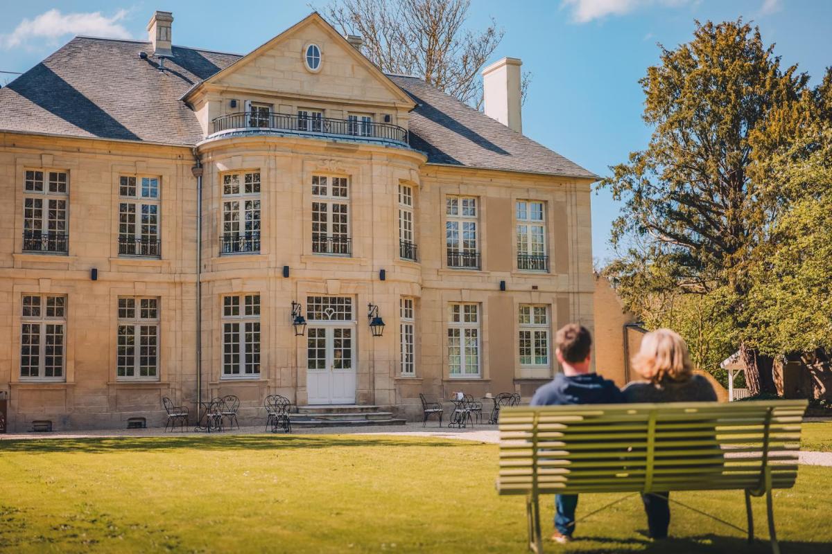 un homme et une femme assis sur un banc devant un grand bâtiment dans l'établissement Domaine de Coeurmandie - Hôtel Château, à Bény-sur-Mer