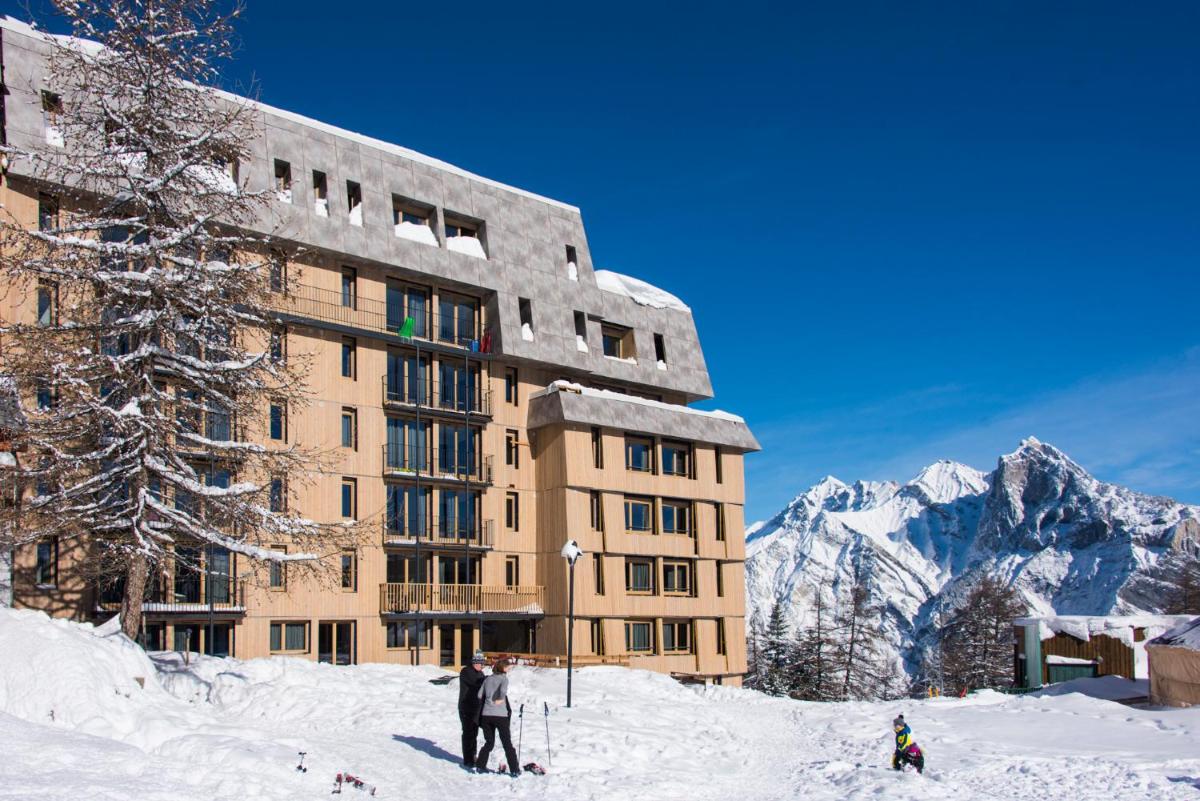 un bâtiment sous la neige avec deux skieurs devant dans l'établissement Résidence Kocoon Les Karellis - Pass activités inclus et accès gratuit et illimité à l'espace aquatique Aquarellis, à Montricher-Albanne