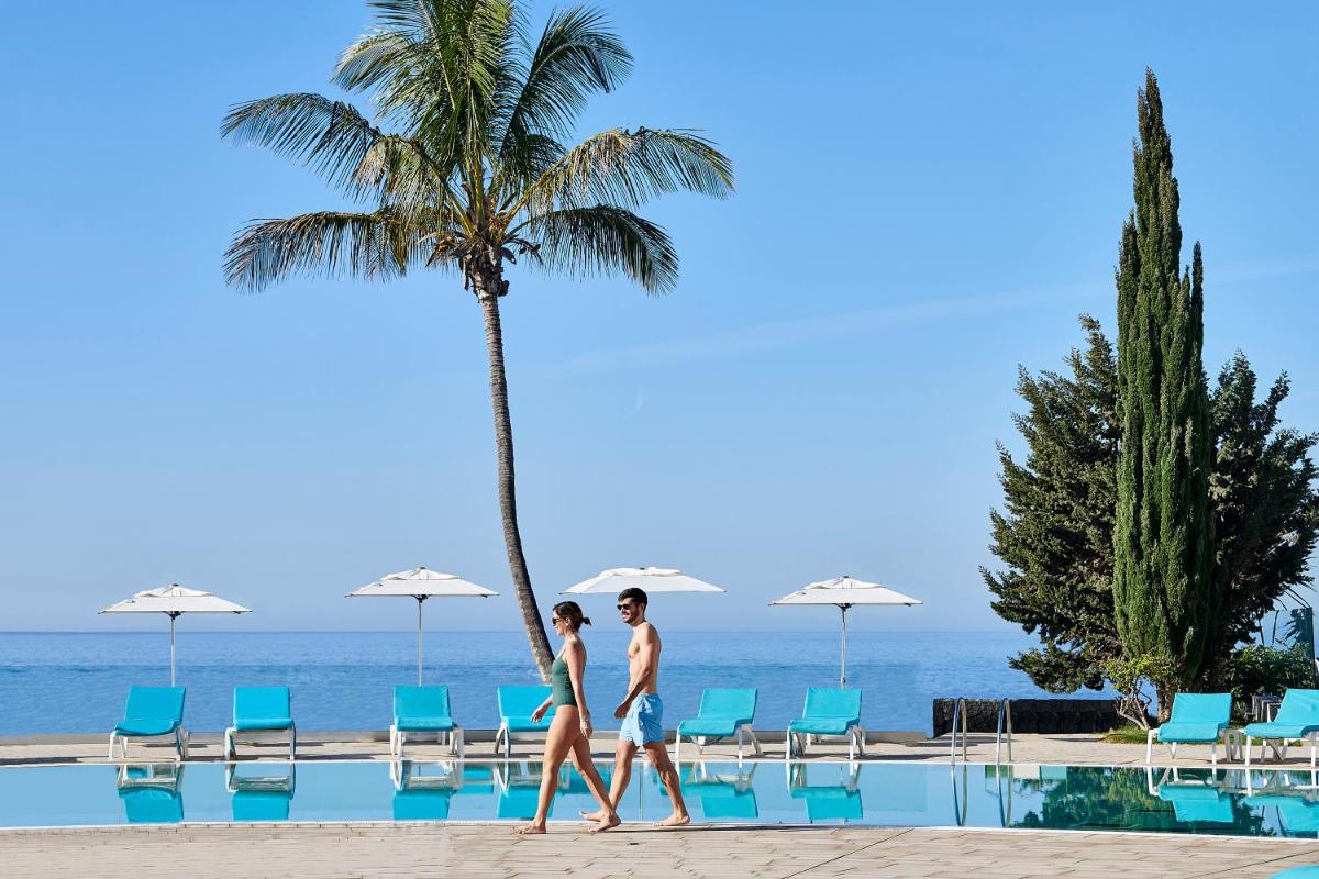 Dos personas caminando junto a una piscina con una palmera. en Iberostar Selection Lanzarote Park, en Playa Blanca