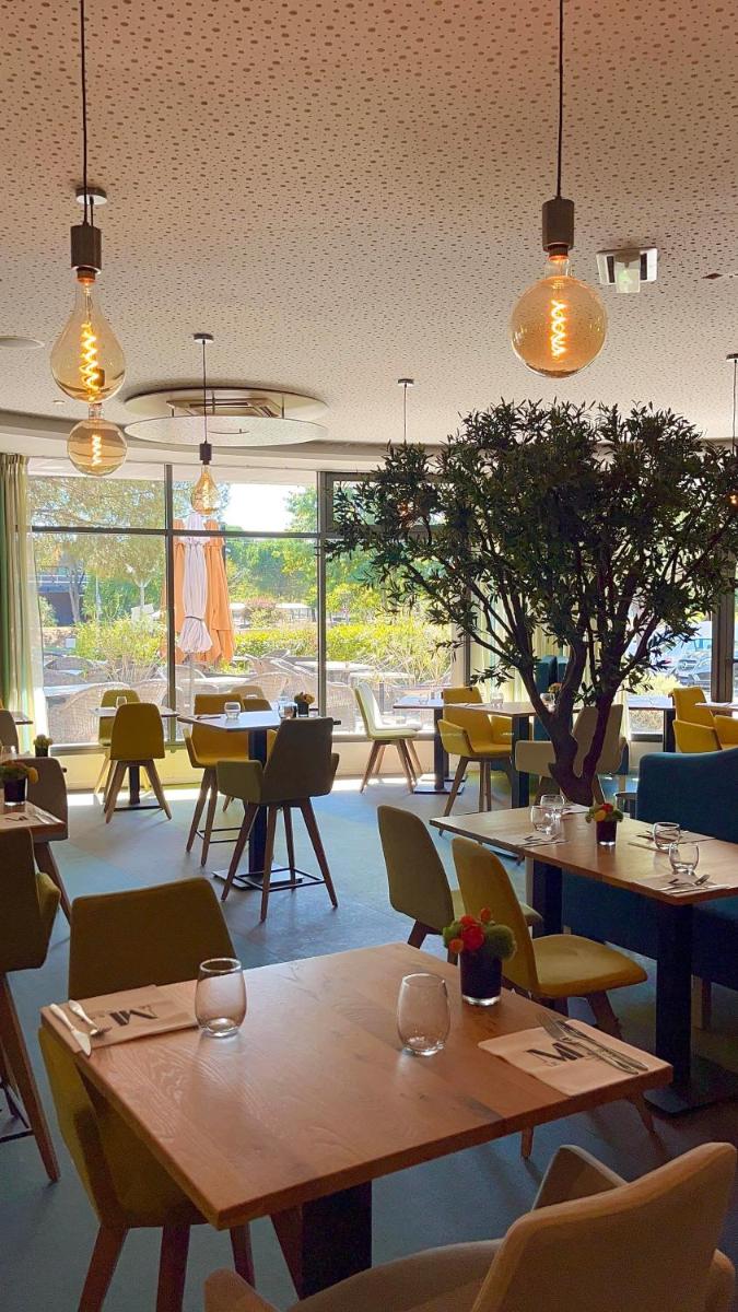 un restaurant avec des tables et des chaises et une grande fenêtre dans l'établissement Le M & Spa Bordeaux Aéroport by Hôtels & Préférence, à Mérignac