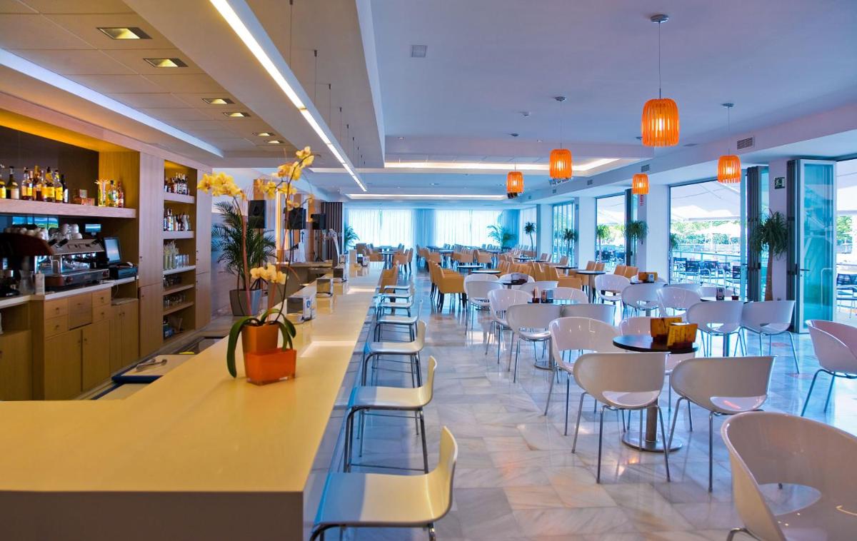 Un restaurante con sillas y mesas blancas y ventanas. en Hotel RH Bayren Parc, en Gandía