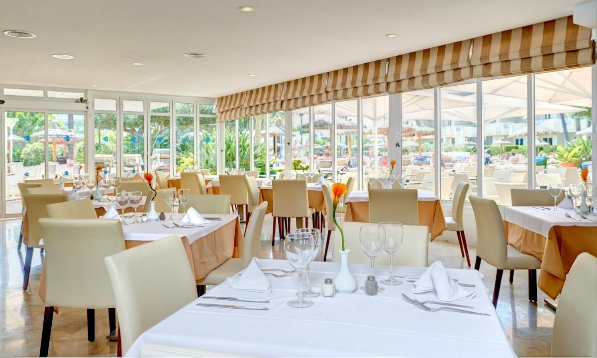 Un restaurante con mesas y sillas blancas y ventanas. en Grupotel Port D´Alcudia, en Puerto de Alcudia