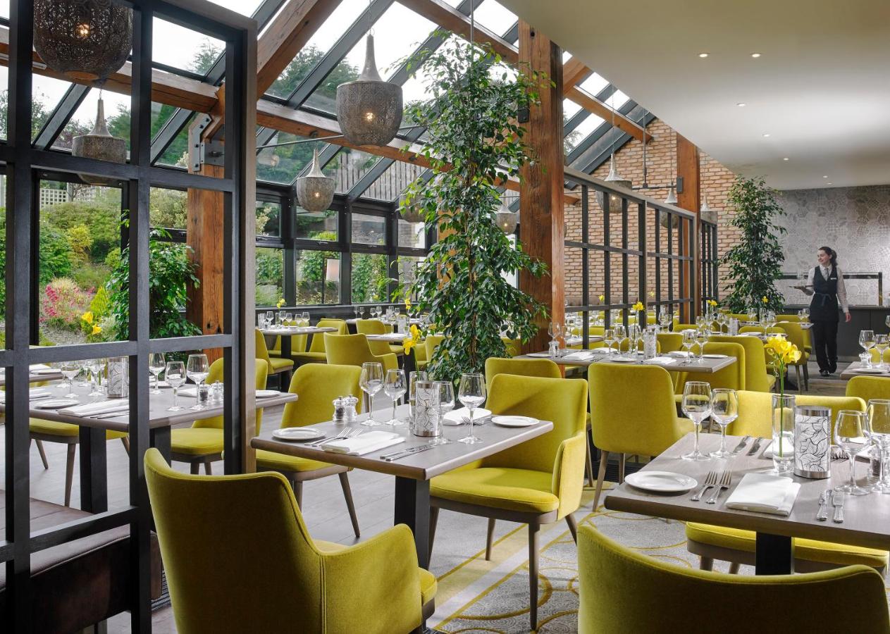 Modern restaurant interior with large windows, yellow chairs, and lush greenery.