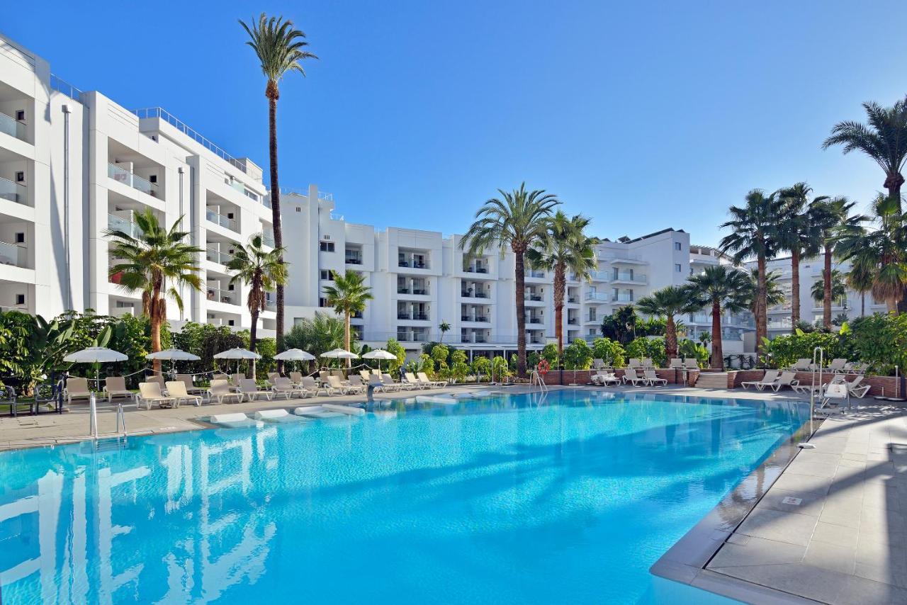 A resort swimming pool flanked by palm trees and loungers with a white building in the background.