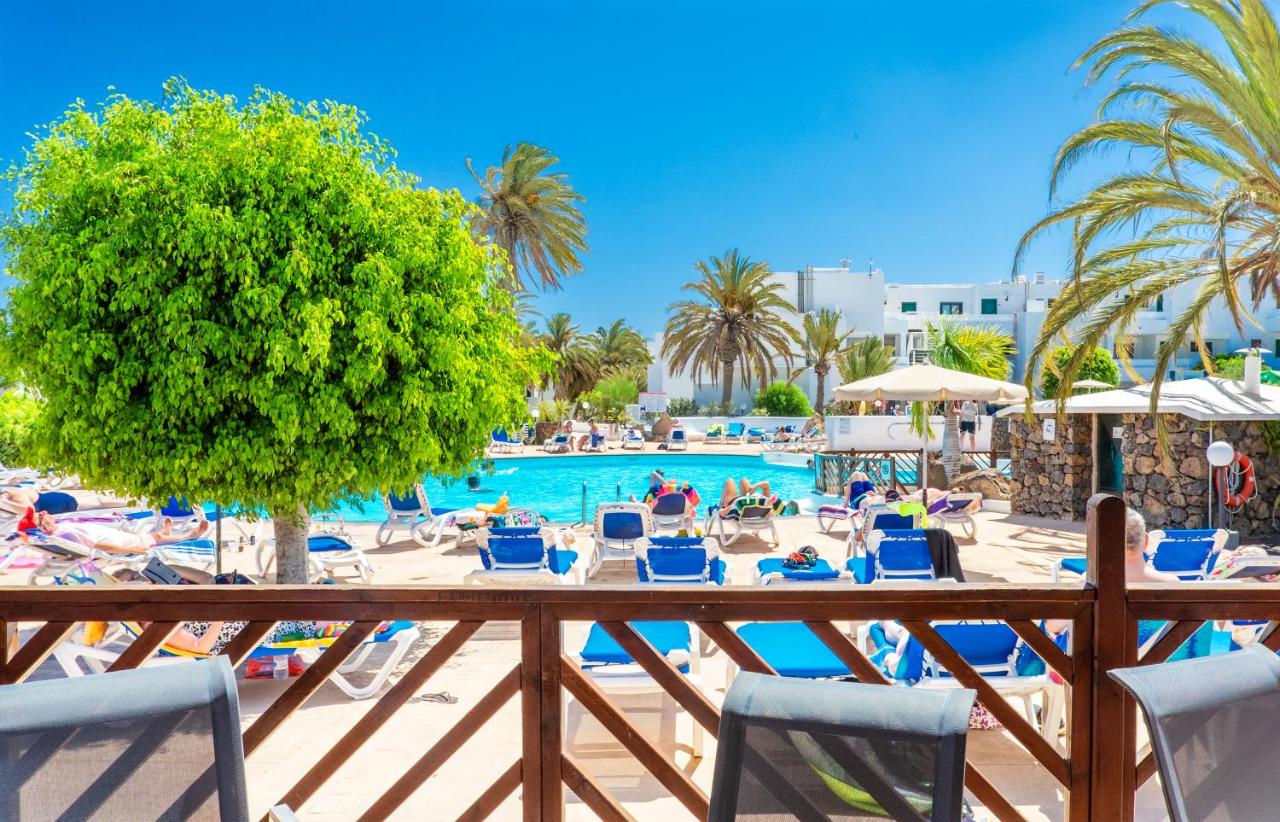 Outdoor resort pool area with sun loungers, umbrellas, and palm trees against a clear blue sky.