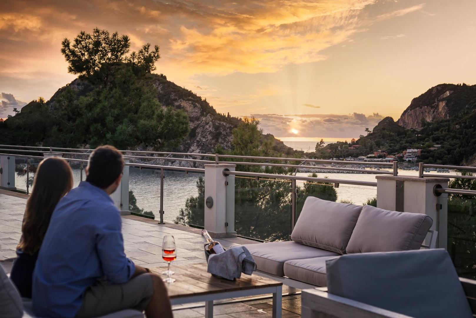 Pāris bauda saulrietu uz terases ar vīna glāzēm un skatu uz kalniem.