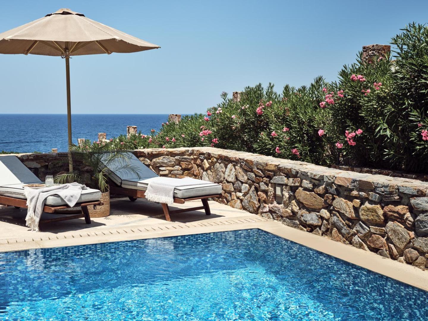 Sauļošanās krēsli un saulessargs pie baseina ar skatu uz jūru un ziedošiem krūmiem.