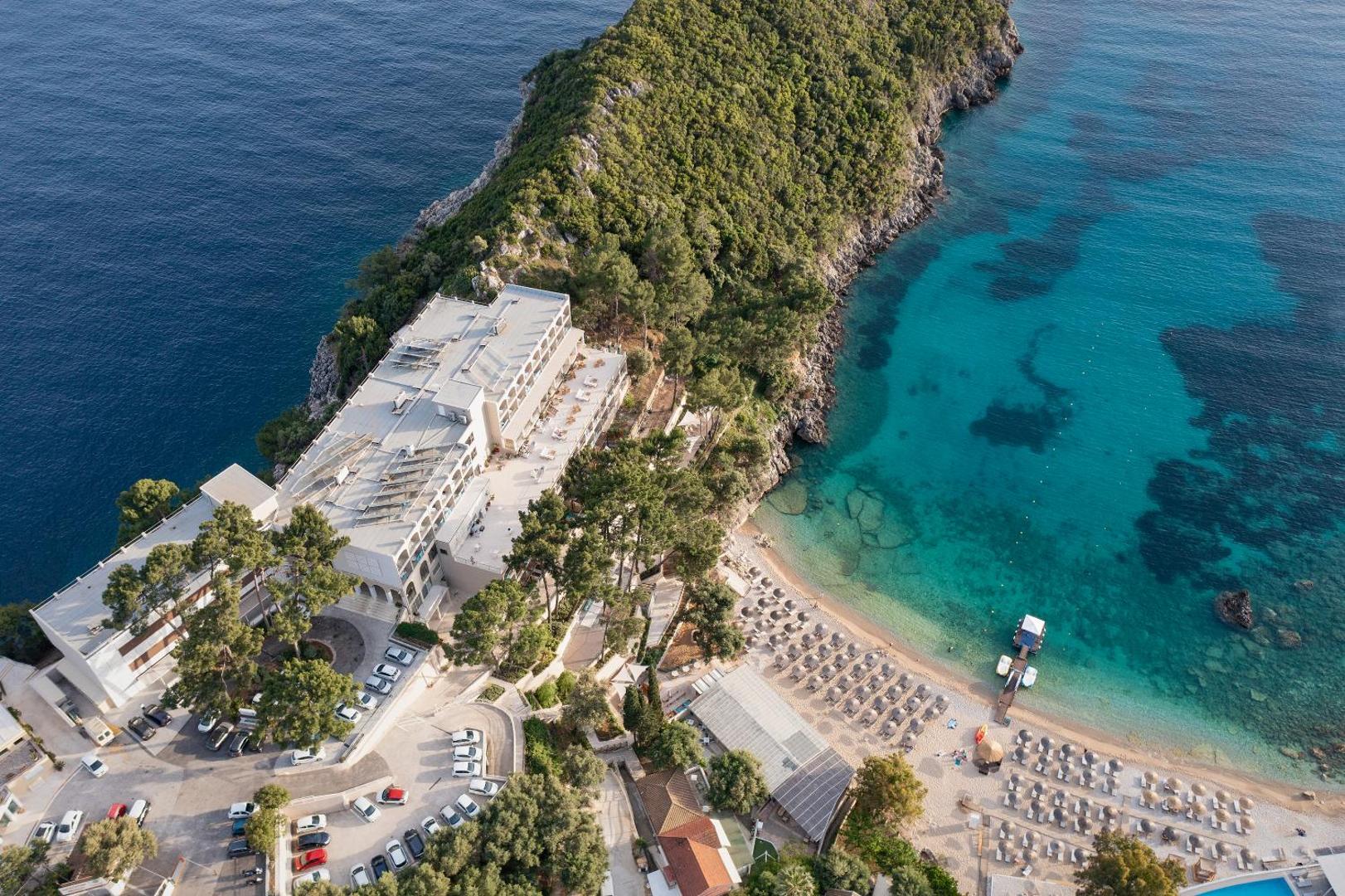 Skats no augšas uz viesnīcas ēku, autostāvvietu un blakus esošo pludmali ar saulessargiem.