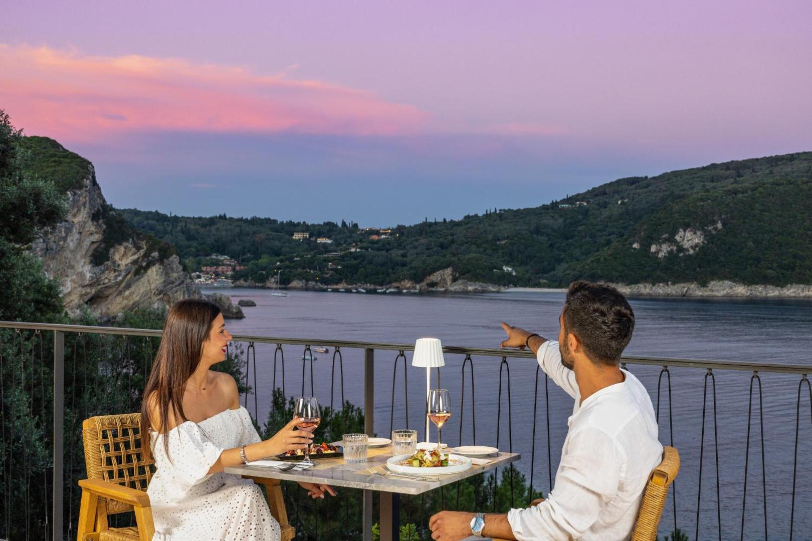 Pāris bauda vakariņas uz balkona ar skatu uz jūras līci saulrieta laikā.