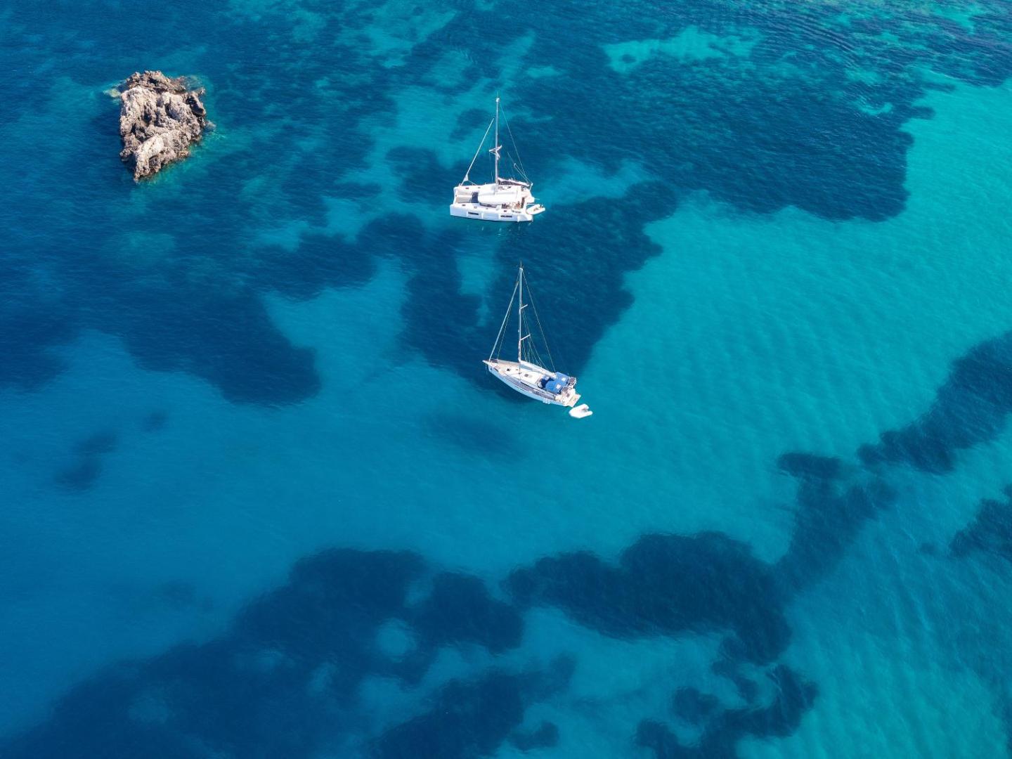 Aerofoto uzņēmums ar divām jahtām kristāldzidrā, tirkīzzilā jūras ūdenī pie klints.