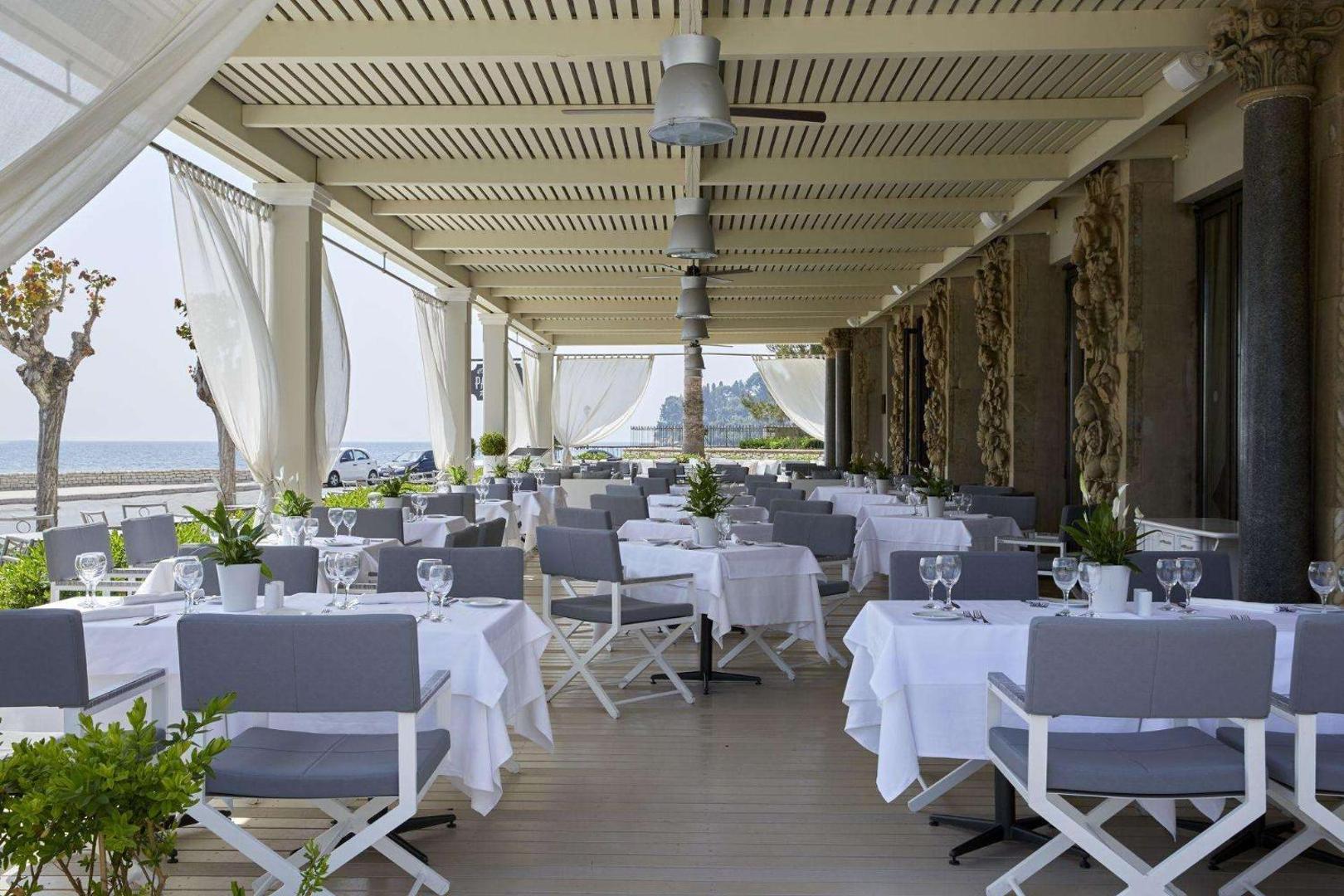 Gaiša un plaša restorāna terase ar pelēkiem krēsliem un skatu uz piekrasti.