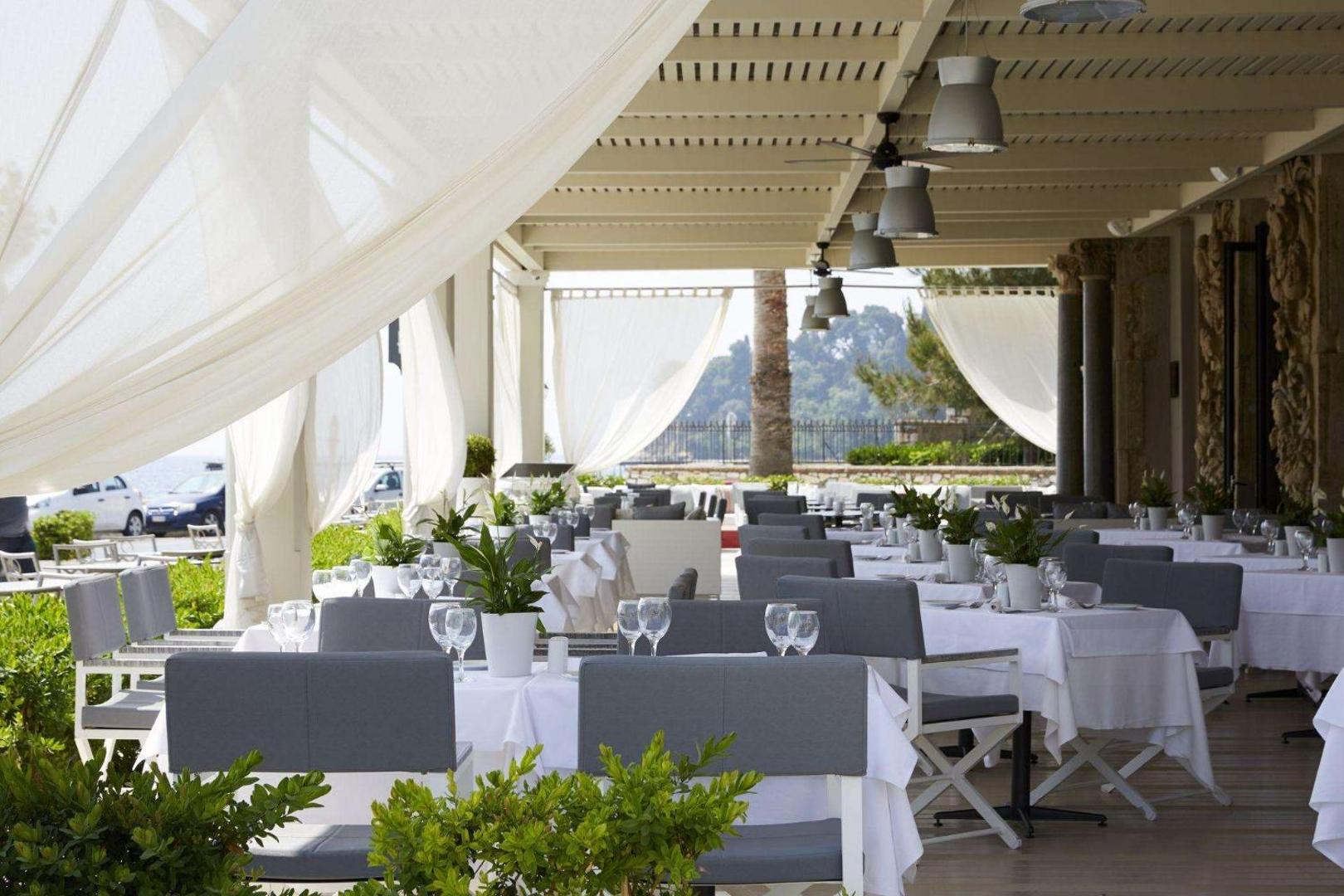 Viesnīcas restorāna āra terase ar eleganti klātiem galdiem un baltiem saulessargiem pie jūras.