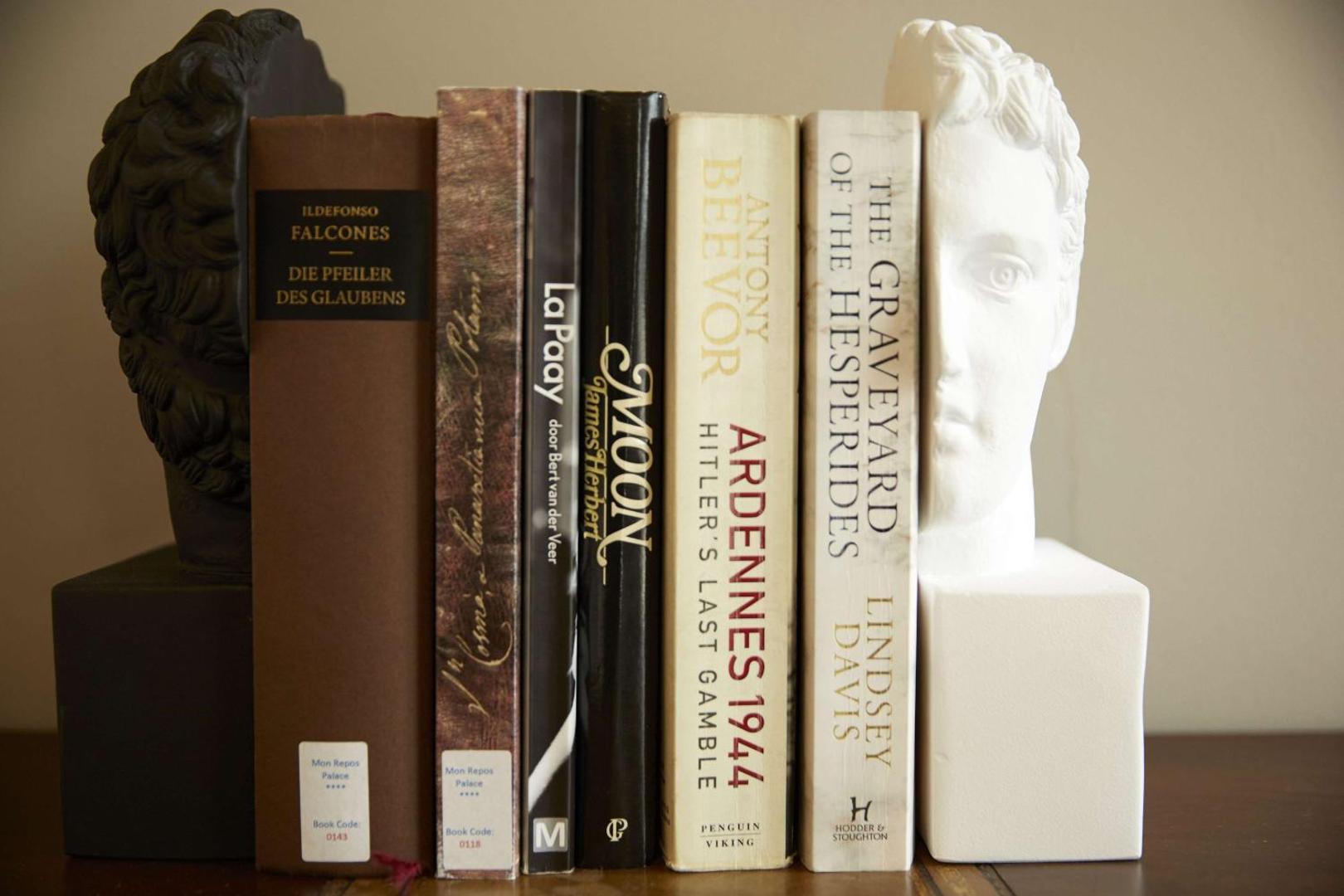 Grāmatu rinda starp diviem antīka stila krūšutēliem uz koka plaukta.