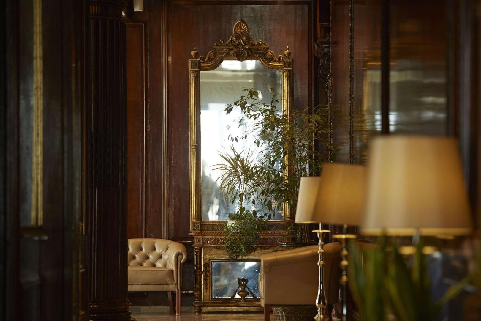Liels, grezns spogulis zeltītā rāmī starp ādas atpūtas krēsliem un telpaugiem.