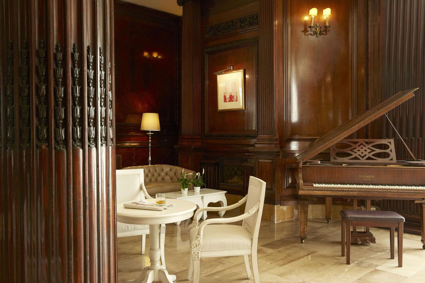 Atpūtas zona ar klavierēm, koka sienu paneļiem un baltiem galdiņiem.