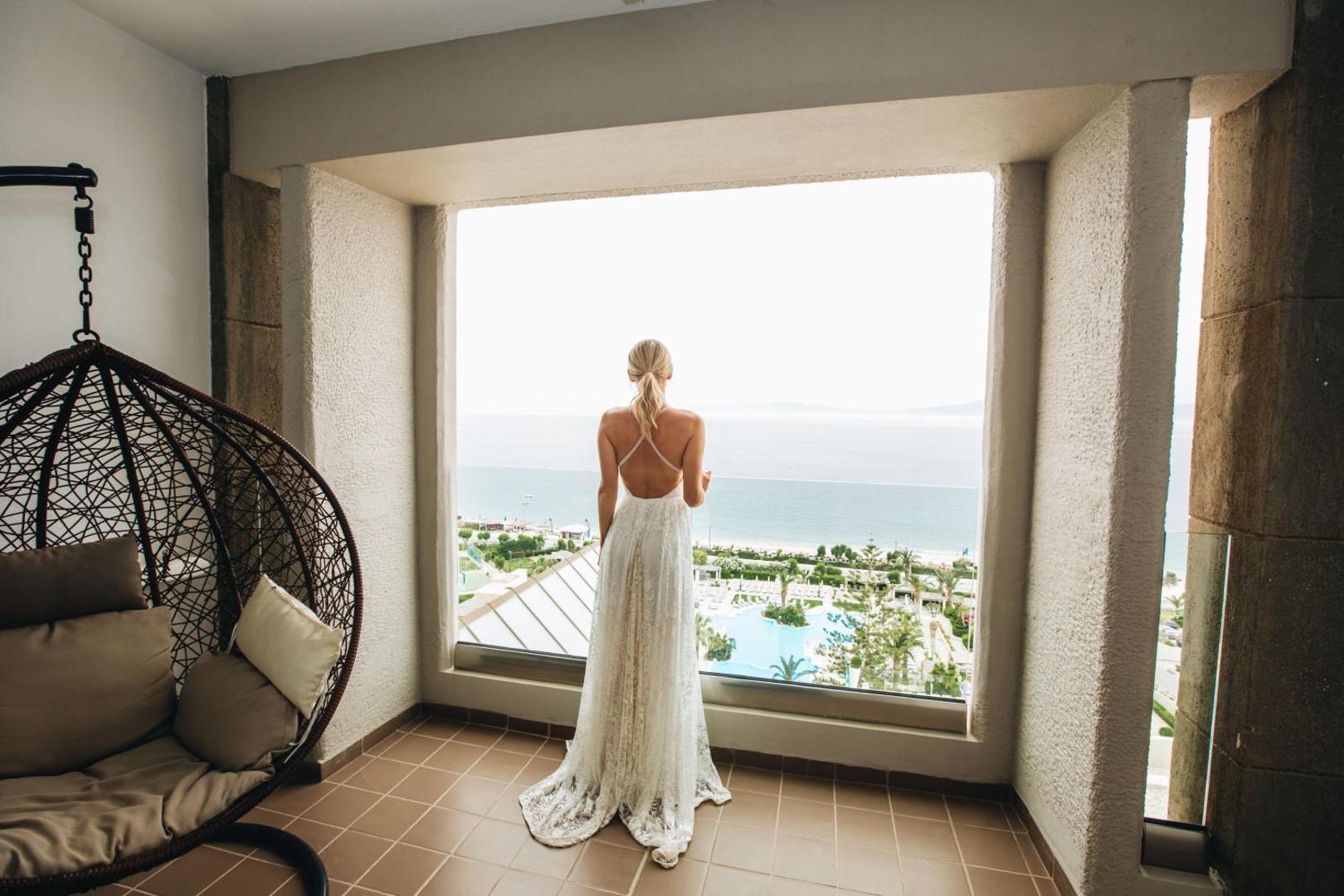 Sieviete baltā kleitā stāv uz balkona, raugoties uz kūrorta baseiniem un jūru.