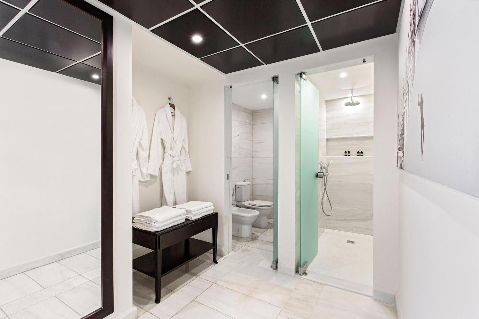 Moderna vannas numura zona ar atsevišķu dušu, tualeti un baltiem peldmēteļiem.