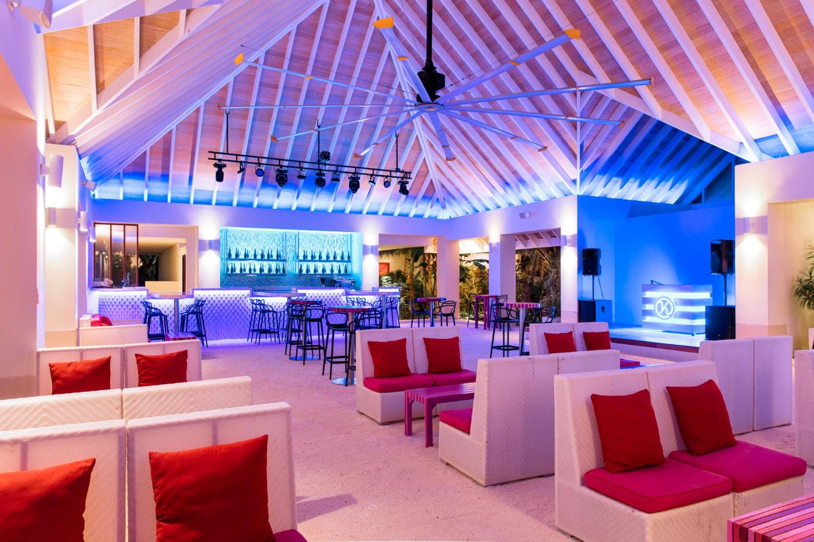 Moderns bārs ar neona apgaismojumu, baltiem dīvāniem un sarkanu akcentu spilveniem.