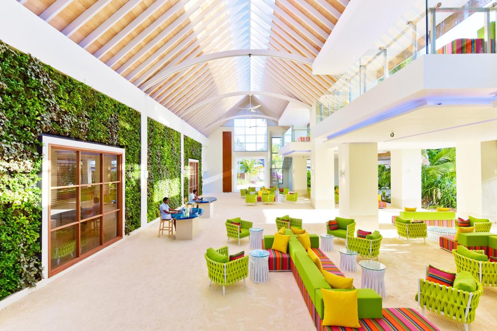 Plašs viesnīcas vestibils ar augstiem griestiem, zaļajām sienām un krāsainām mēbelēm.