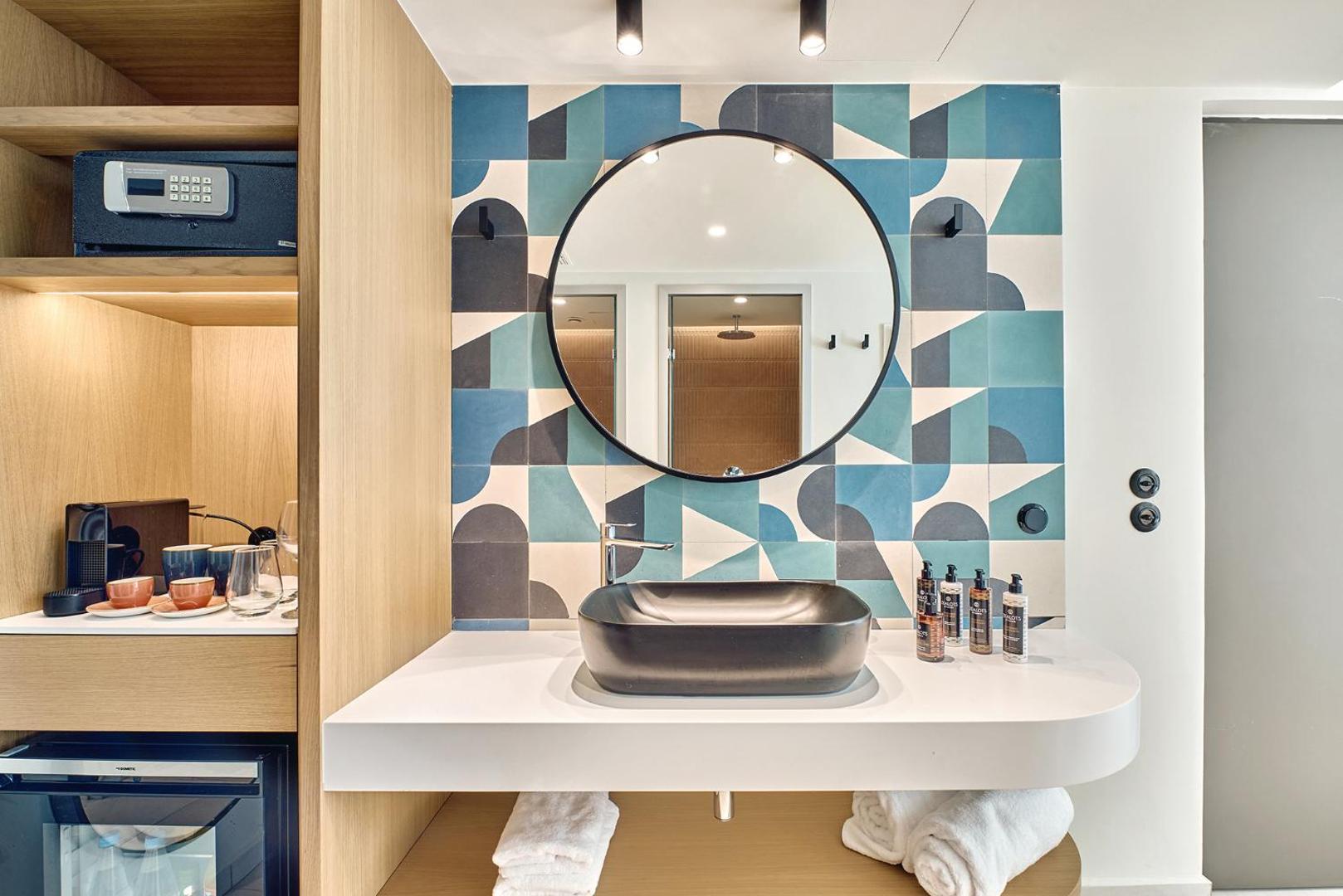 Mūsdienīgs viesnīcas numura interjers ar melnu izlietni, apaļu spoguli un zilu rakstainu flīžu sienu.