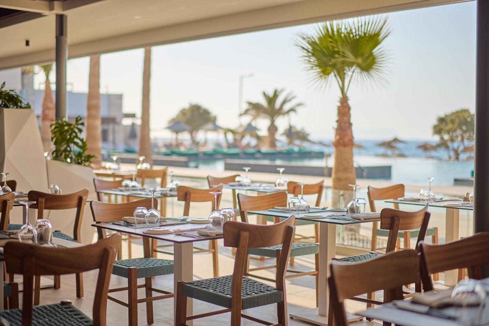 Eleganti klāti galdi viesnīcas restorāna terasē ar skatu uz baseinu un palmu.