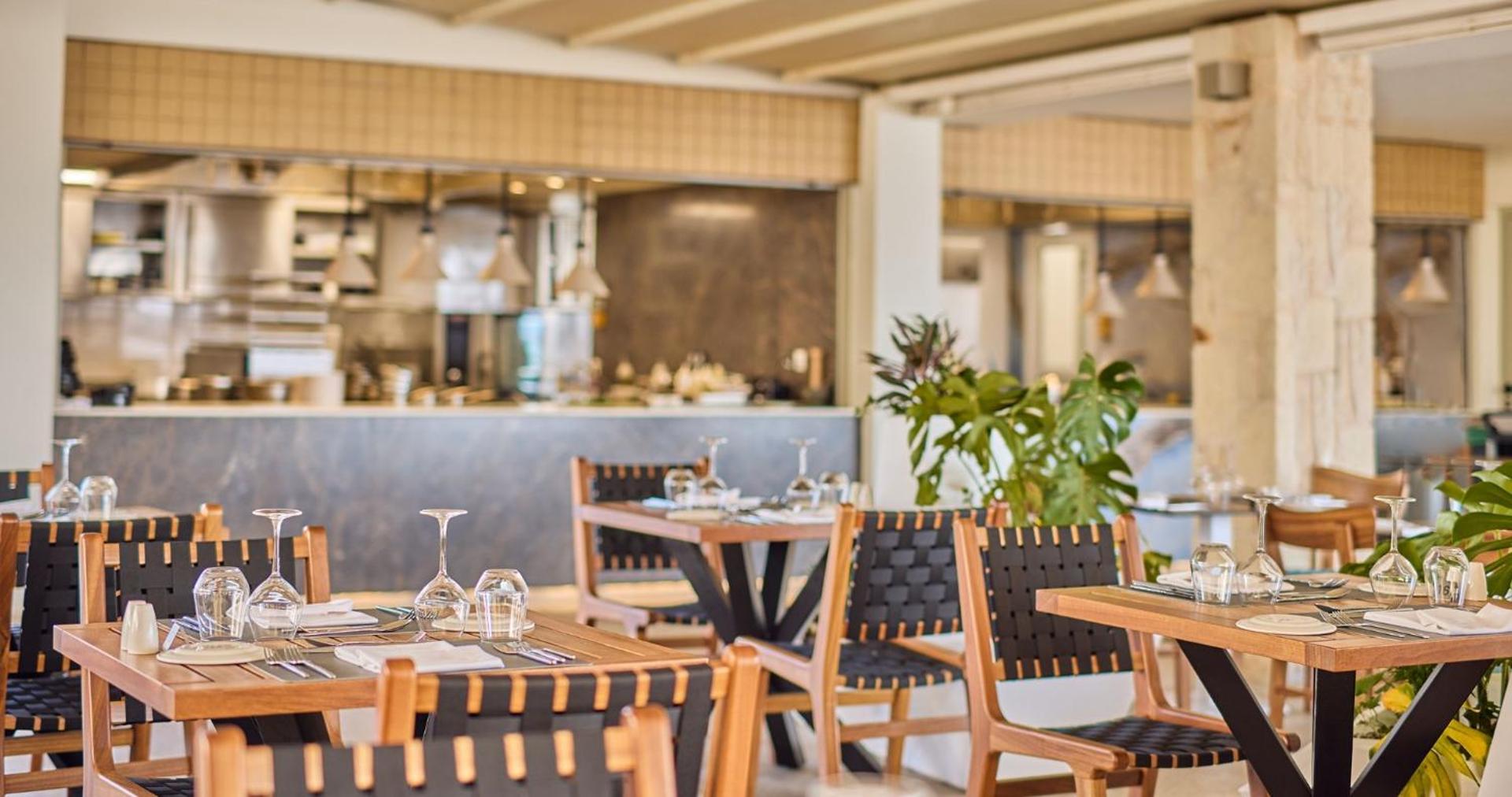 Gaišs viesnīcas restorāns ar koka galdiem un pītiem krēsliem mājīgā atmosfērā.