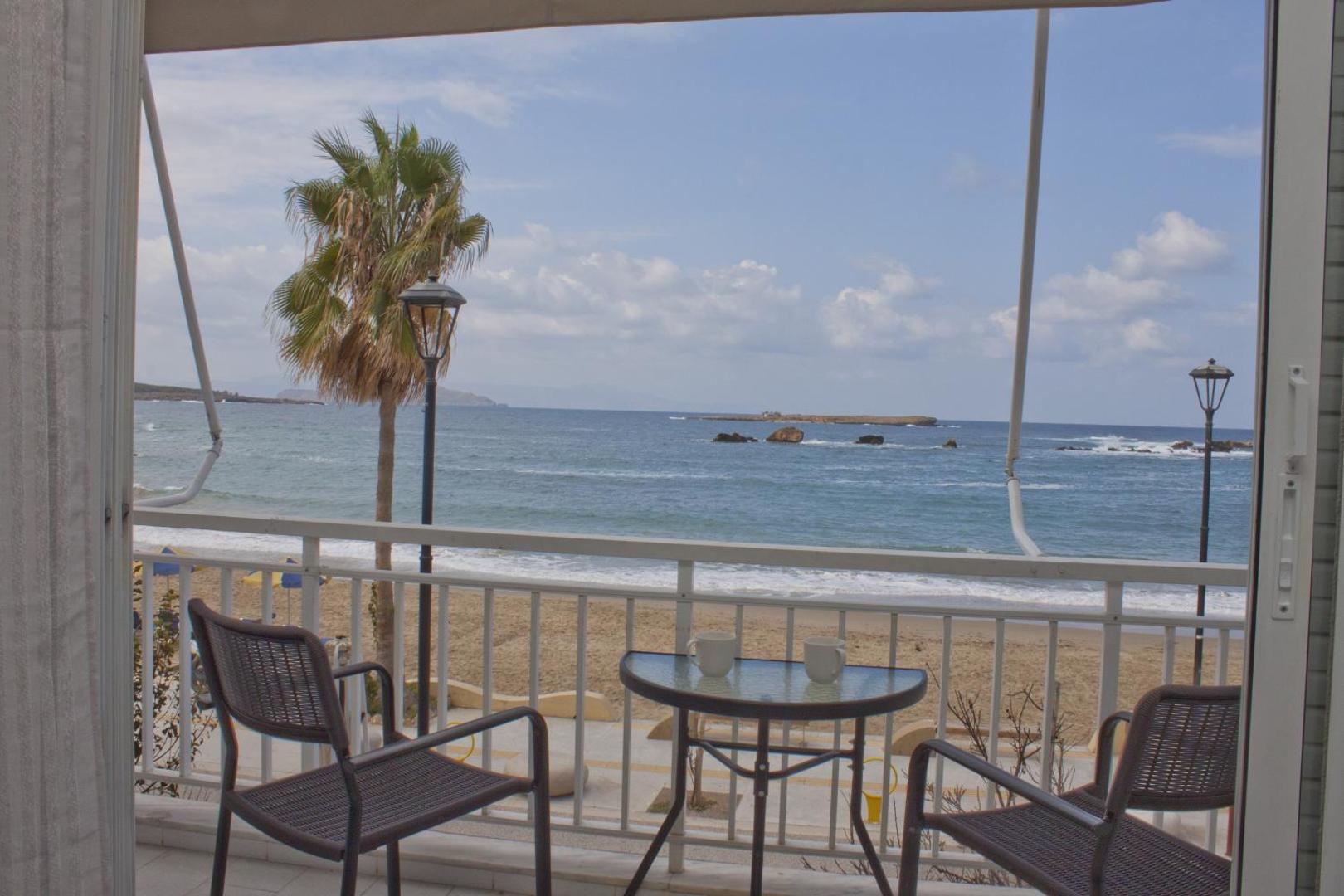 Skats caur atvērtām balkona durvīm uz galdiņu, krēsliem un jūras panorāmu.