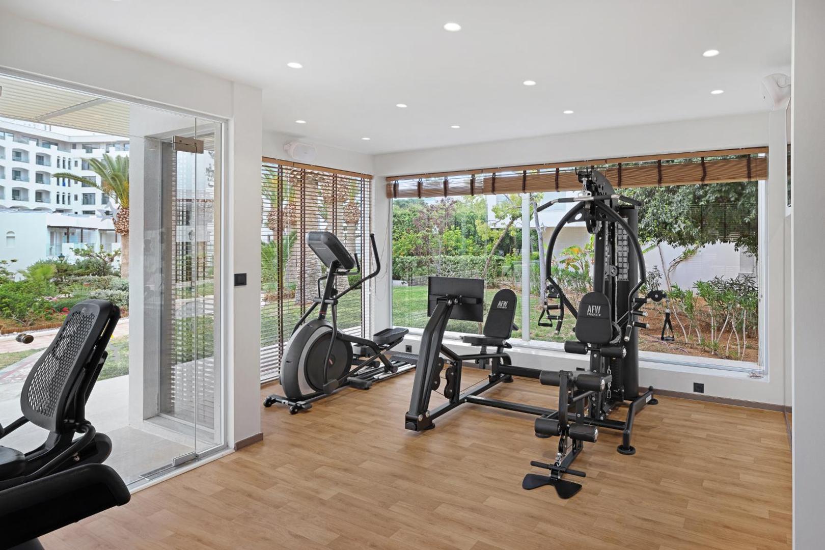 Fitnesa zāle ar spēka trenažieriem un lieliem logiem uz zaļo zonu.