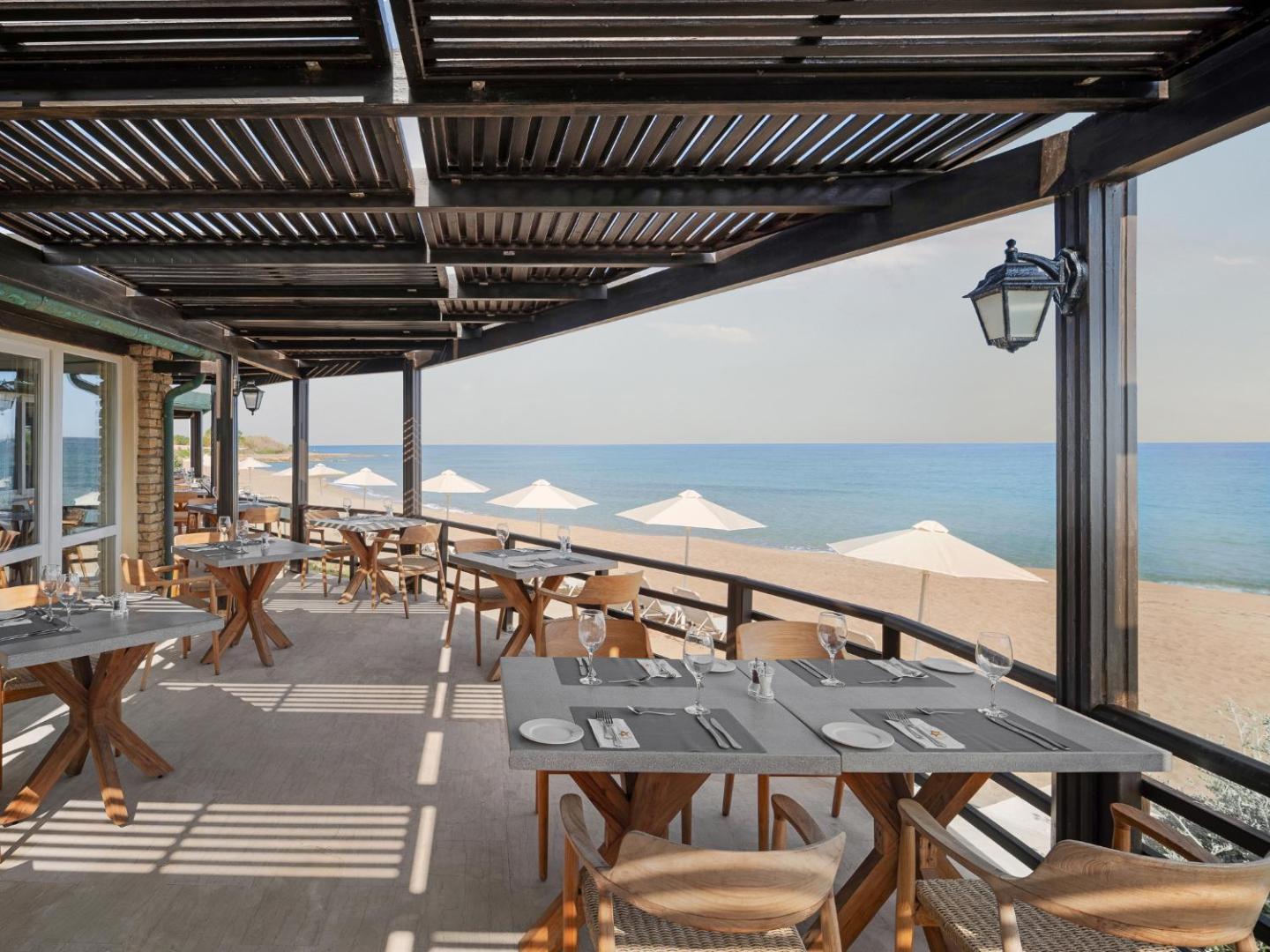Restorāna terase ar koka galdiem un krēsliem, no kuras paveras skats uz pludmali.