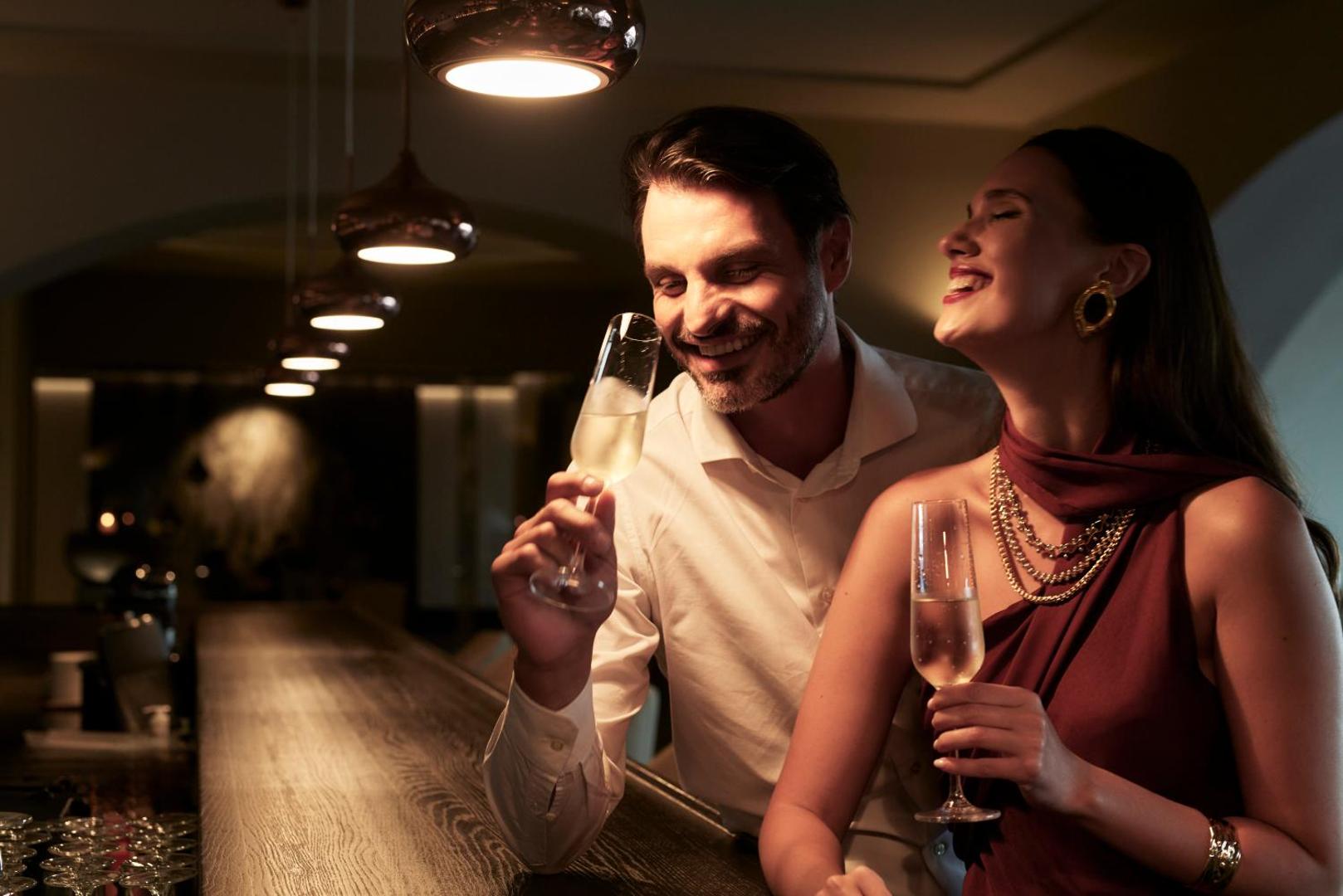 Smaidīgs pāris bauda dzirkstošo vīnu pie viesnīcas bāra letes vakara apgaismojumā.